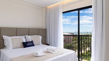 A hotel room with a large bed, white bedding, a headboard, and a view of the beach through a balcony.