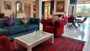Een ruime woonkamer met een blauwe bank, een rode fauteuil en een glazen salontafel. Er is een eethoek met rode stoelen en een kamerbreed tapijt met een patroon.