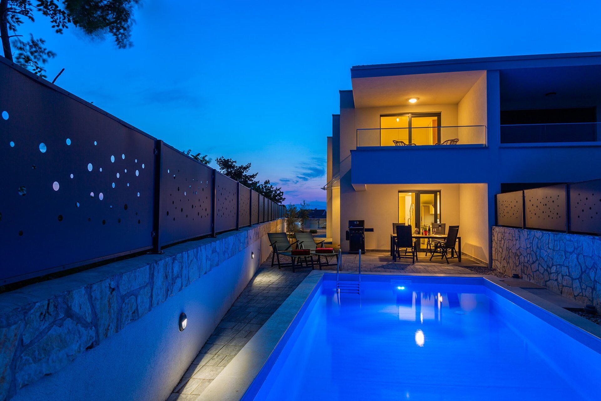 A modern two-story building with a swimming pool, outdoor seating, and a stone wall.