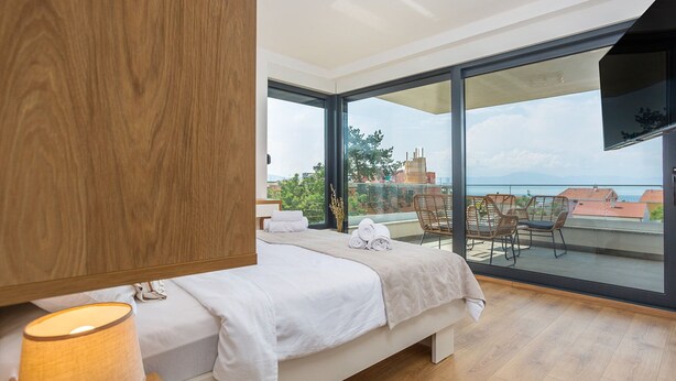 A bedroom with a bed, a wooden wardrobe, a bedside lamp, and a balcony with a view of the sea and buildings. - Villa Felice with Pool (Malinska)