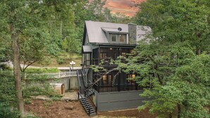 A two-story cabin with a stone foundation, a wooden deck, and a staircase leading to the upper level. - Ultimate Creek Retreat- private 250 ft waterfront - Hot Tub, Sauna, Game Room (Epworth)