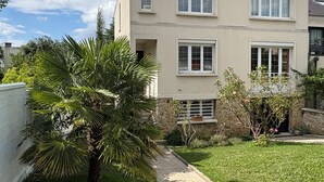 A two-story building with a flat roof, multiple windows, and a small garden area with a palm tree and shrubs. - A HAVEN OF PEACE ON THE OUTSKIRTS OF PARIS (Clamart)