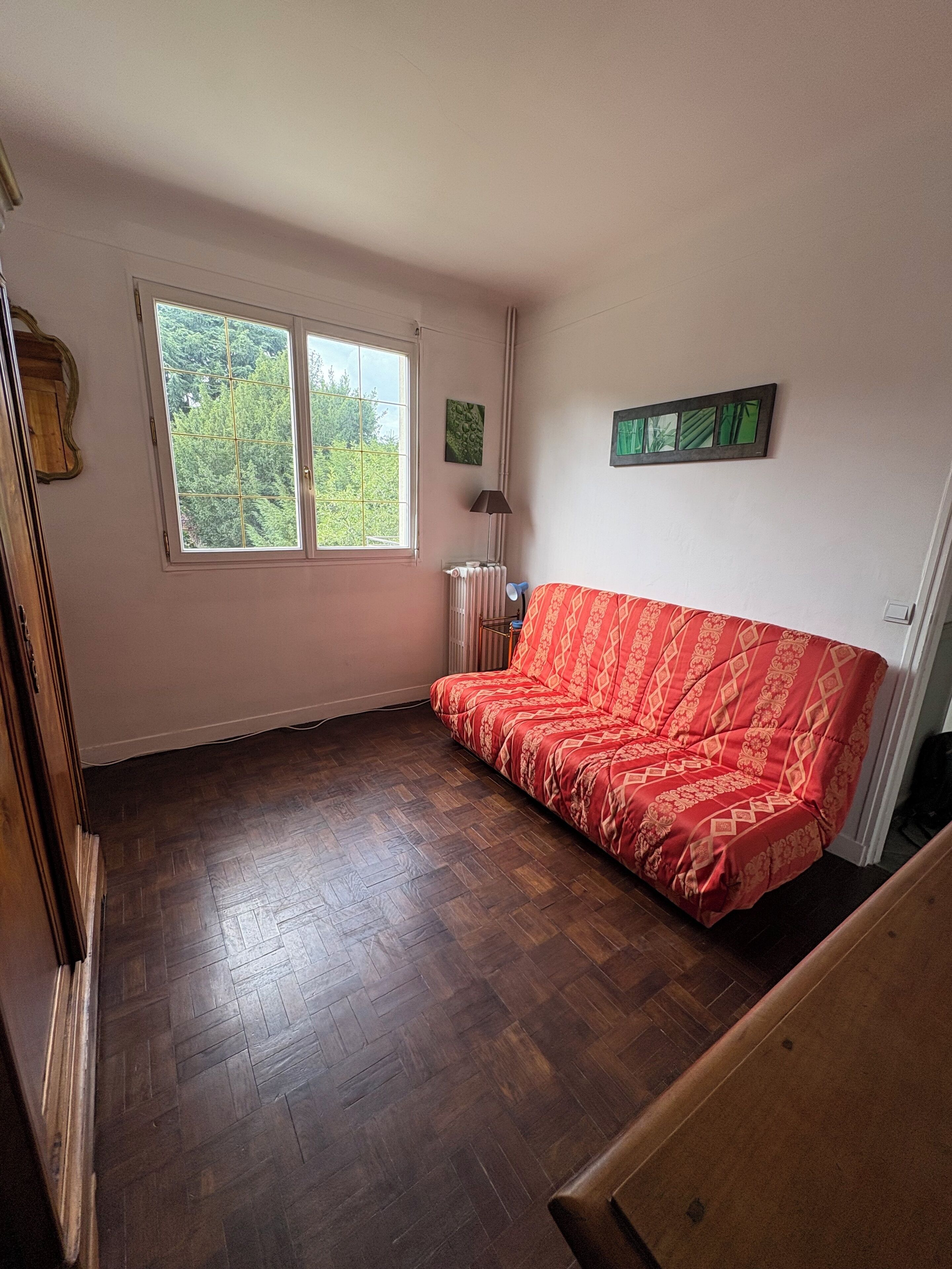Une pièce avec un plancher en bois, un canapé rouge, une fenêtre donnant sur un paysage verdoyant, un radiateur et une lampe.