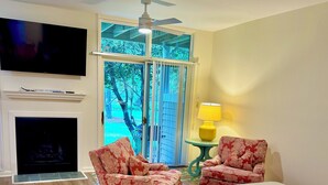 A living room with a fireplace, a television, a ceiling fan, and a view through a large window. - Cozy Lakeview Stay by the Beach (Rehoboth Beach)