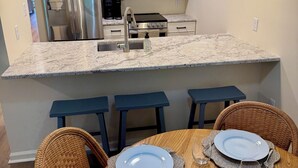 A modern kitchen with white cabinets, stainless steel appliances, and a marble countertop. There are four blue bar stools and a dining table set with plates and glasses. - Cozy Lakeview Stay by the Beach (Rehoboth Beach)
