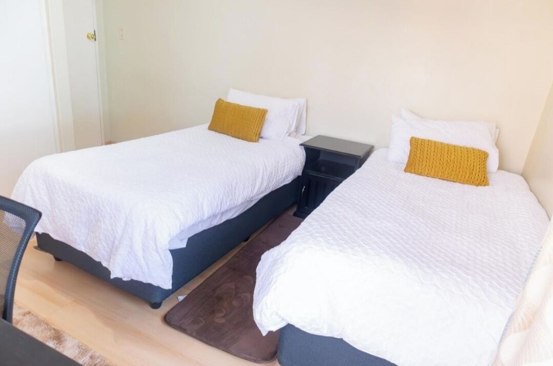 Dos camas individuales con ropa blanca y almohadas color mostaza, una mesita y una silla en una habitación sencilla con suelo de tonos claros.