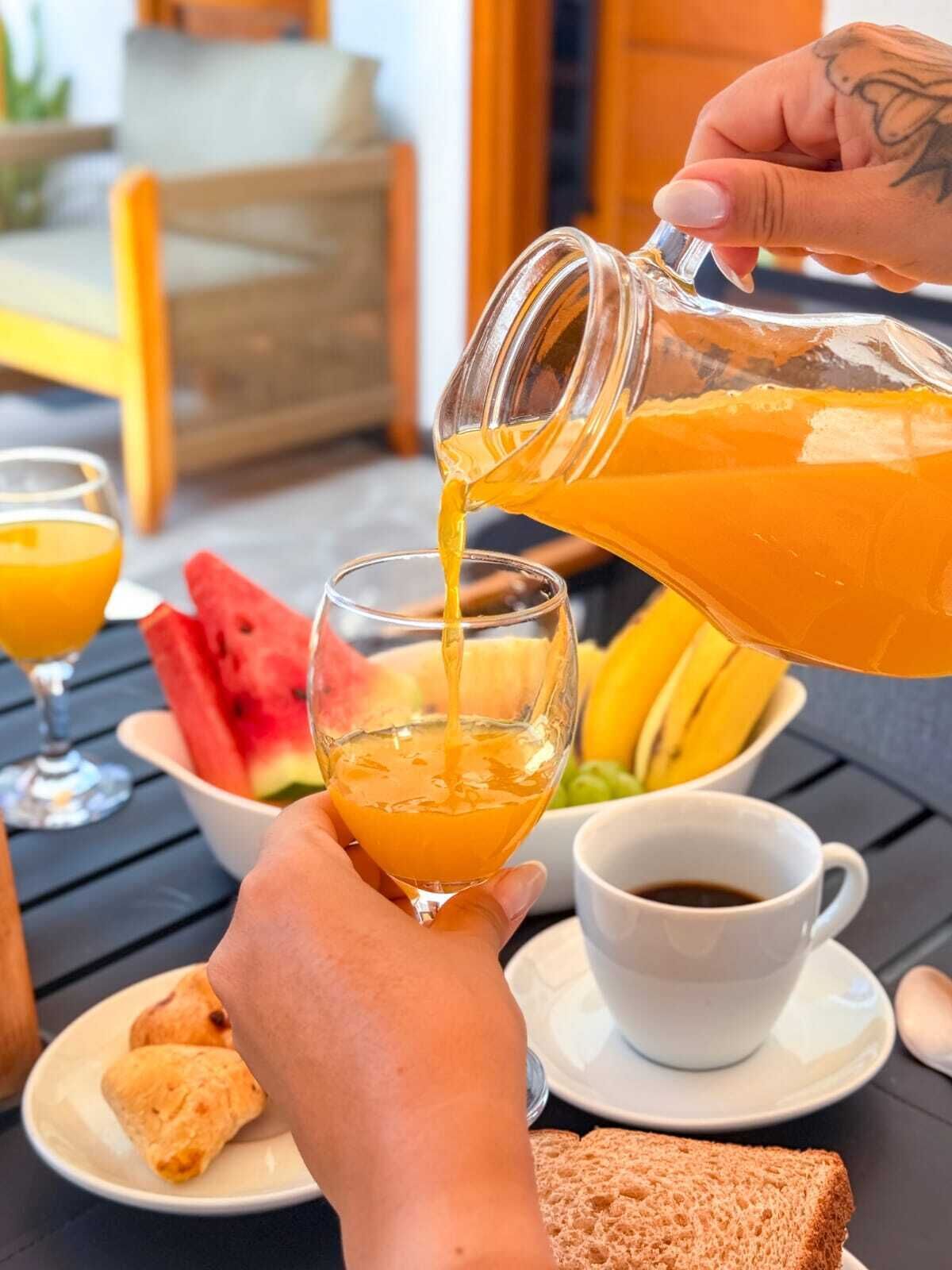 A breakfast setting with a person pouring orange juice into a glass, a bowl of watermelon, bananas, and toast on the table.