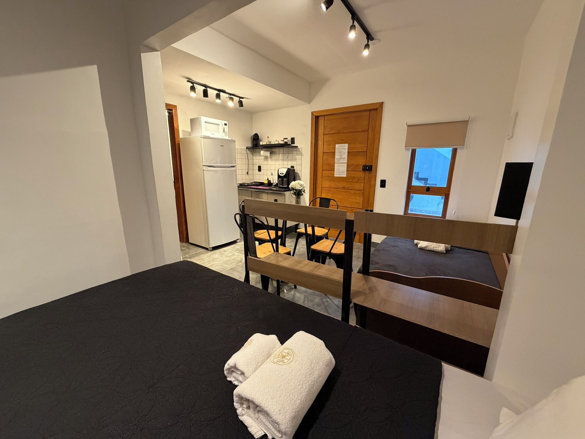 A bedroom with a bed, a bench, and a view of a kitchen and bathroom.