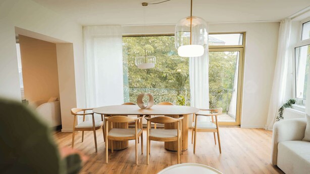 A dining area with a round table and six chairs, a white coffee table with a book, and large windows with a view of greenery.