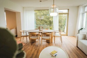 A dining area with a round table and six chairs, a white coffee table with a book, and large windows with a view of greenery.