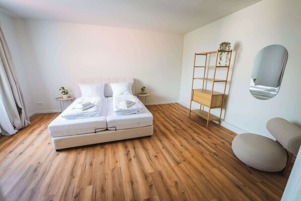 A modern bedroom with a large bed, a wooden floor, a small side table with a plant, a mirror, and a wooden shelf unit.