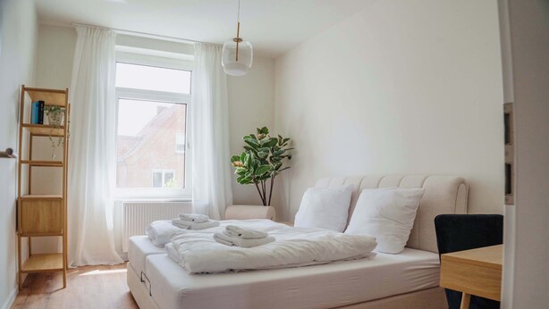 Un dormitorio moderno con una cama grande, una mesita de noche de madera, un escritorio pequeño, una estantería y una planta en maceta.