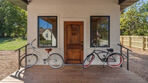A wooden porch with two bicycles parked in front of a white building with a wooden door and two windows. - Missouri’s 1st Sinclair Service Station (Walnut Grove)