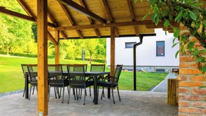 An outdoor patio with a wooden pergola, a table with chairs, and a brick pathway leading to a building.