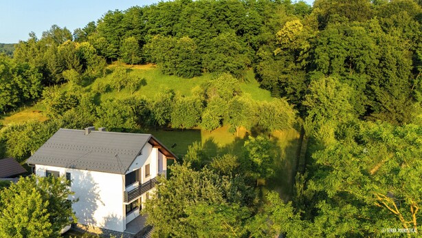 A modern two-story house with a sloped roof, surrounded by dense forest and a small clearing.