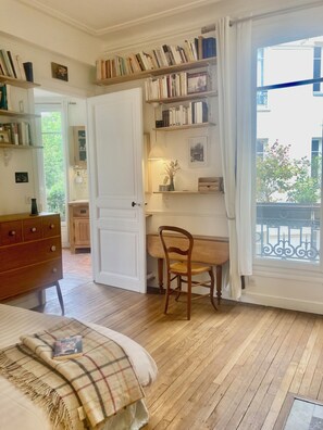 Ein gemütliches Zimmer mit einem Holztisch, einem Stuhl, einem Bett mit karierter Decke und einem Bücherregal voller Bücher.