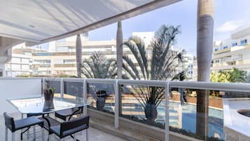 A balcony with a glass table and chairs, a potted plant, and a view of a swimming pool and buildings.