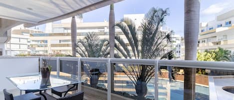 Un balcón con una mesa y sillas de vidrio, una planta en maceta y vistas a una piscina y edificios.
