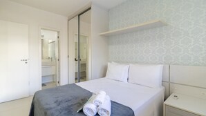 A neatly made bed with a grey bedspread and rolled white towels, a bedside table with a power outlet, and a wardrobe with a mirror. - Apart w/ Barbecue in Condominium With Pool Bri0102 (Florianópolis)