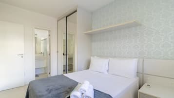 A neatly made bed with a grey bedspread and rolled white towels, a bedside table with a power outlet, and a wardrobe with a mirror.