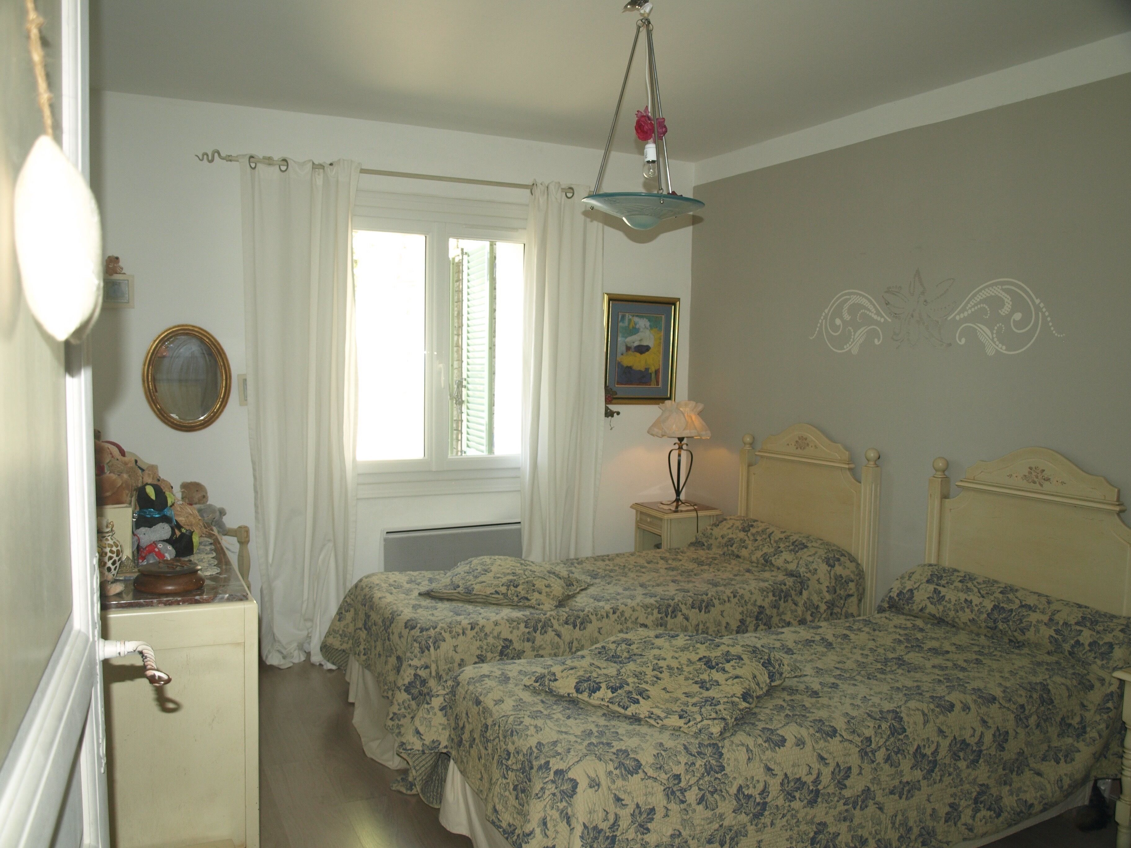 Une chambre avec deux lits, une commode, un miroir, une fenêtre avec des rideaux, un radiateur, une lampe et un tableau encadré au mur.