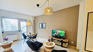 Una sala moderna con televisor, una mesa de comedor con sillas, una mesita de mimbre y un amplio ventanal con vistas a edificios.