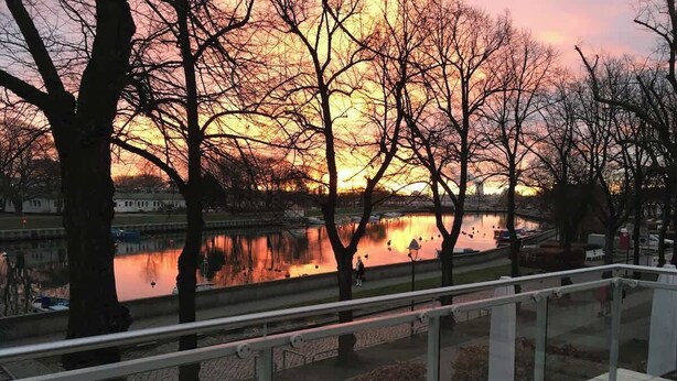A riverside walkway with a railing, trees, and a sunset in the background. - Helle Wohnung mit Balkon am Strom in Warnemünde (Rostock)