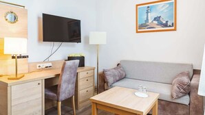 A hotel room with a wooden desk, a grey sofa, a TV mounted on the wall, a painting of a lighthouse, and a bedside lamp. - Krusenstern (Rostock)