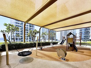 Zona de juegos al aire libre con columpios, tobogán y estructura de escalada, rodeada de vegetación y edificios modernos al fondo.