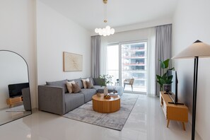 A modern living room with a grey sofa, a round wooden coffee table, a TV on a wooden stand, and a large window with a view of buildings.