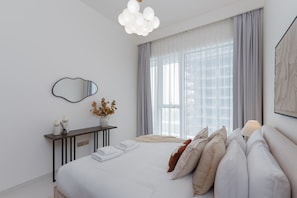 Un dormitorio moderno con una cama grande, una mesita de noche con un florero, un espejo y una ventana con cortinas.
