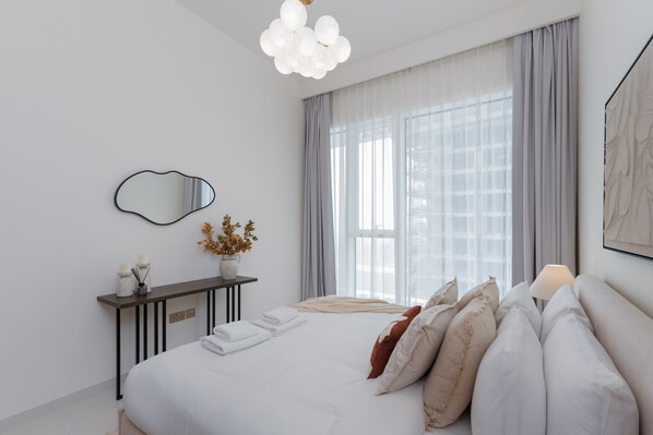 Un dormitorio moderno con una cama grande, una mesita de noche con un florero, un espejo y una ventana con cortinas.