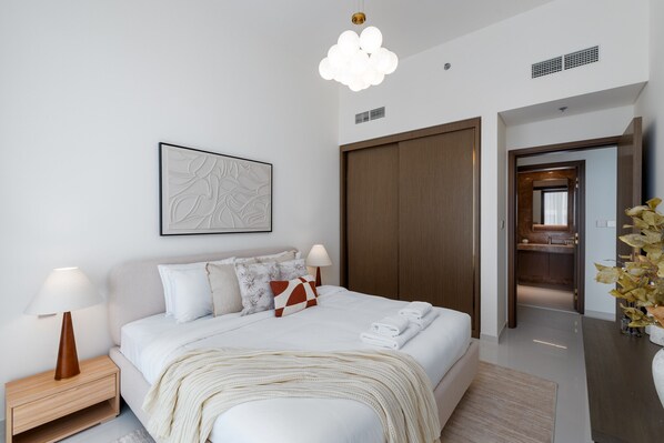 A modern hotel room with a large bed, bedside tables with lamps, a wooden wardrobe, a bathroom visible through an open door, and a decorative plant.