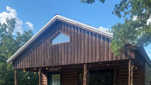 A rustic log cabin with a green door, a covered porch with two rocking chairs, and a small window above the door.