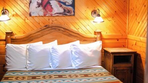 A wooden cabin room with a bed, two wall-mounted lamps, a ceiling fan, and a painting of horses.