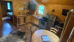 A cozy cabin interior with a wooden ceiling, a dining area with a table and chairs, a kitchen with modern appliances, and a large fur rug on the floor.