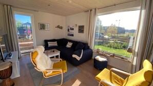 Ein gemütliches Wohnzimmer mit einem dunkelblauen Sofa, gelben Sesseln und einem kleinen Tisch. Ein großes Fenster bietet Blick auf eine grüne Landschaft.