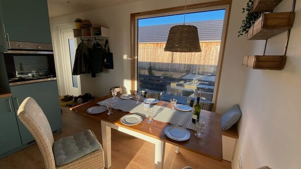 Ein Essbereich mit einem Holztisch, gedeckt für vier Personen, einem Korbstuhl und Blick auf einen Garten durch ein großes Fenster.
