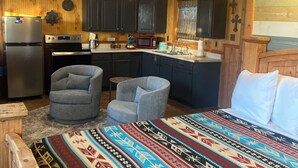 A compact kitchen with dark cabinets, a refrigerator, and a stove. There are two grey armchairs and a small table. A colorful rug is on the floor.