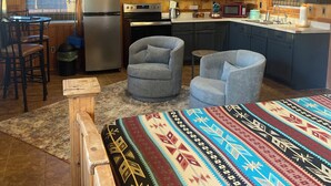 A rustic cabin interior with a kitchenette, two armchairs, a dining table, and a large bed with a patterned blanket.