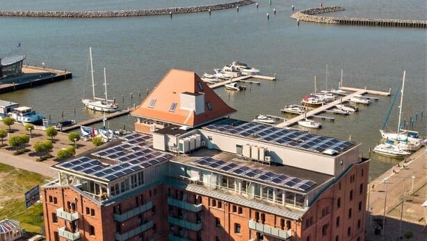A multi-story building with a rooftop covered in solar panels, situated near a marina with boats docked.