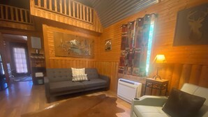 L’intérieur chaleureux d’un chalet, avec un revêtement mural en bois, un canapé gris, un fauteuil blanc, une grande moquette et un tableau accroché au mur.