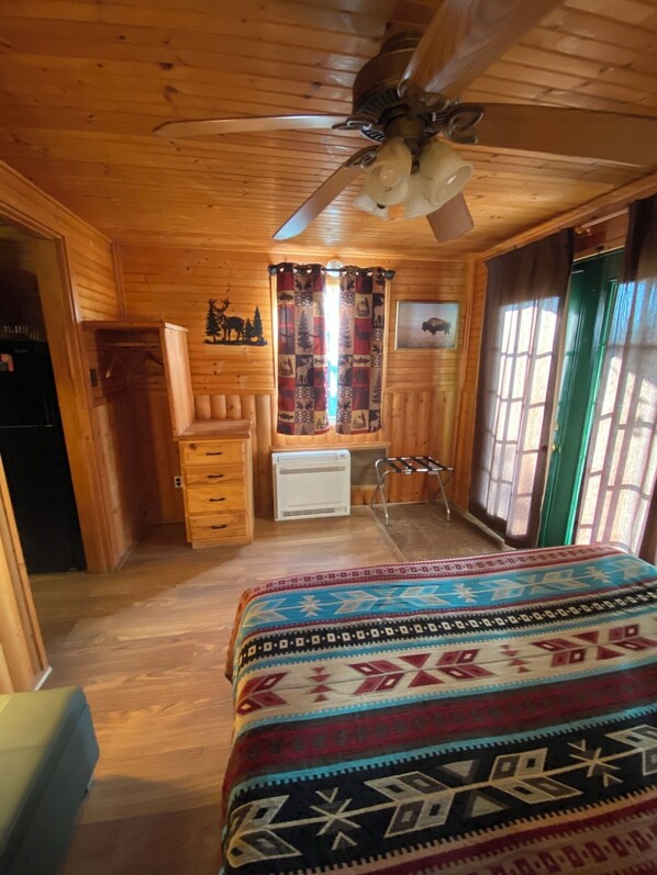 A wooden cabin room with a bed, a ceiling fan, a radiator, and a window with curtains.
