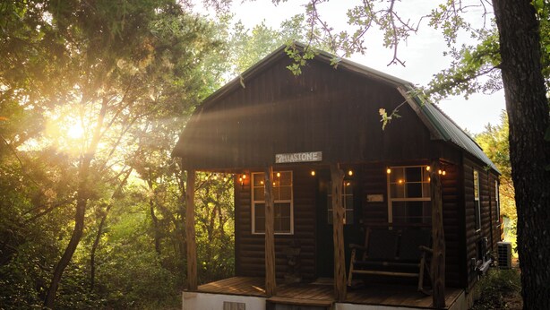 Une cabane rustique en rondins de bois, dotée d’un porche et portant l’inscription « Yellestone », est entourée d’une forêt dense.