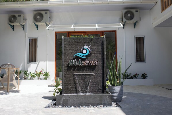 A welcome sign with a wave design, air conditioning units, and potted plants in front of a building.