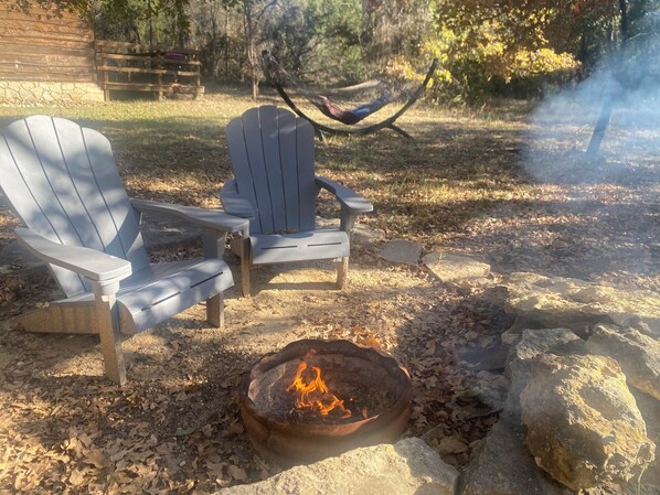 A fire pit with a fire, two Adirondack chairs, and a hammock in a wooded outdoor setting.