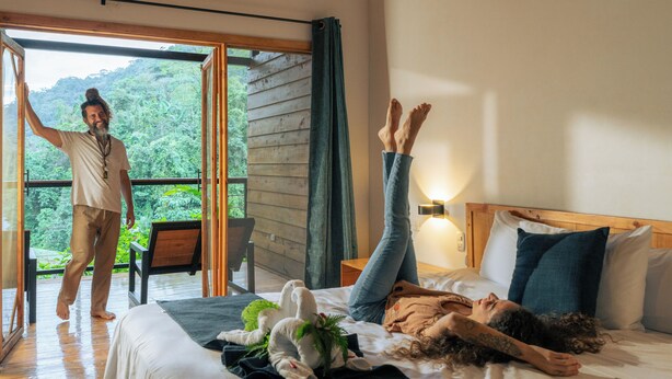 A bedroom with a bed, a person lying on the bed, and another person standing by a sliding glass door. - RIOS LODGE (Pacuarito)