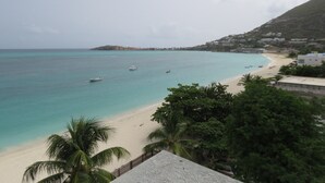 A coastal beach with clear turquoise waters, a sandy shore, and a small hill with buildings in the background. - Charming 2bdrm/2bath beachfront PH Condo WiFi, AC, TV, beach lounges included (Philipsburg)