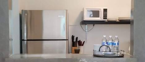 Een functionele keuken met een roestvrijstalen koelkast, een magnetron en een klein plankje met keukengerei en waterflessen. Er staan twee metalen barkrukken met houten zitjes.