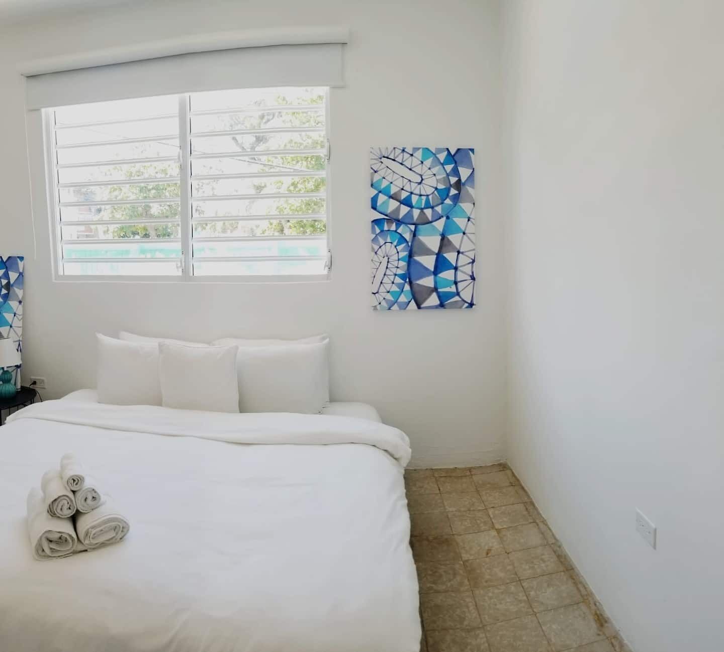 Una habitación de hotel con una cama, una ventana con persianas y una obra de arte geométrica en la pared.